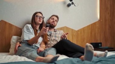 Genç çift otel odasında televizyon izliyor ve gülümsüyor.