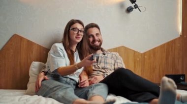 Genç çift otel odasında televizyon izliyor ve gülümsüyor.