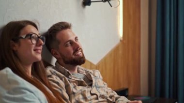 Genç çift otel odasında televizyon izliyor ve gülümsüyor.