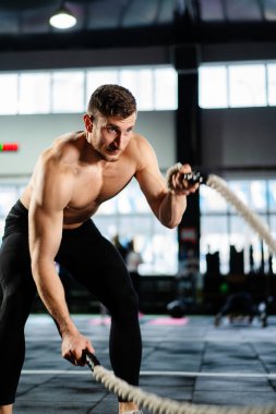 Koyu renk spor salonunda güçlü bir adam dövüş ipleri eğitiyor. Profesyonel atlet spor salonu ekipmanları. Güçlü vücut geliştirici ağırlık kaldırıyor. Atletik kişi çabası