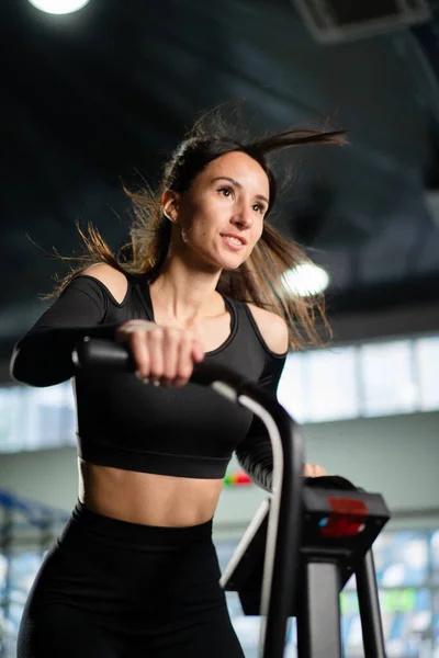 Young Athletic middle aged woman training on air resistance bike, cross training workout set in gym. Active woman spinning a air bike in gym with trainers. female training on air bike.