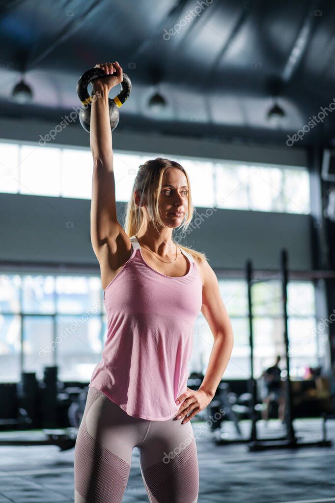 a woman lifts a dumbbell over her head while working out in a dark gym. Athletic strong woman performs a difficult workout with lifting dumbbells