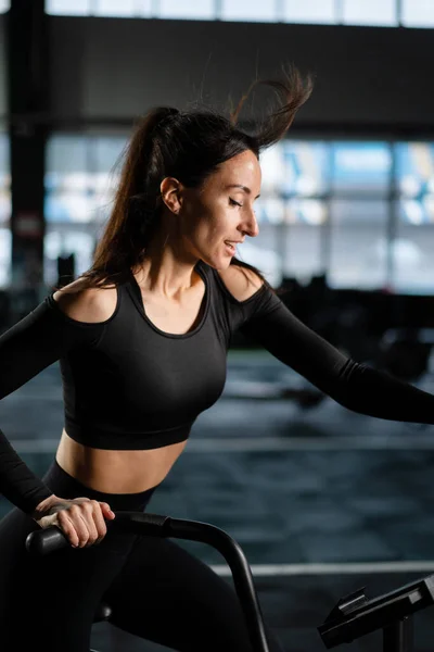 Young Athletic middle aged woman training on air resistance bike, cross training workout set in gym. Active woman spinning a air bike in gym with trainers. female training on air bike.