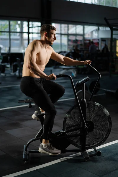 Young Athletic middle aged man training on air resistance bike, cross training workout set in gym. Active man spinning a air bike in gym with trainers. Male training on air bike.