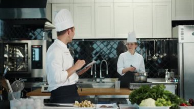 Restaurant kitchen: portrait of male and female cooks. Male cook checks the availability of products in the warehouse using a tablet