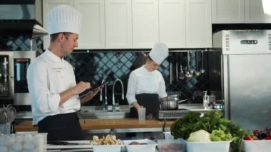 Restaurant kitchen: portrait of male and female cooks. Male cook checks the availability of products in the warehouse using a tablet