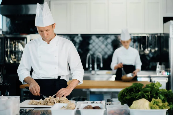 Professional kitchen: two chefs prepare food. The cook cuts mushrooms to prepare delicious dish.