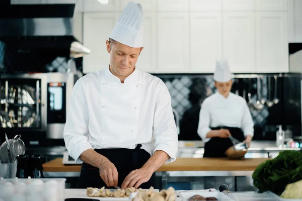 Professional kitchen: two chefs prepare food. The cook cuts mushrooms to prepare delicious dish.