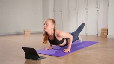 Determined athletic woman exercising, stretching exercises on yoga mat while watching online fitness course video on tablet. Woman practicing yoga online with laptop