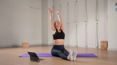Determined athletic woman exercising, stretching exercises on yoga mat while watching online fitness course video on tablet. Woman practicing yoga online with laptop