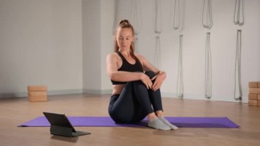 Determined athletic woman exercising, stretching exercises on yoga mat while watching online fitness course video on tablet. Woman practicing yoga online with laptop