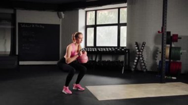 Attractive young beautiful woman doing cardio exercise and squatting with kettlebell in gym