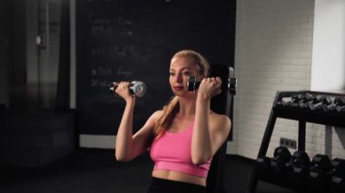 The girl does an exercise in gym. Female athletic woman exercising shoulders with dumbbells