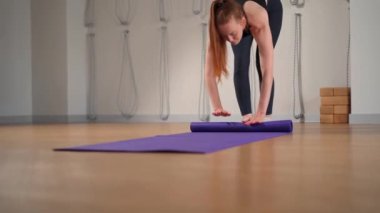 Woman rolling up yoga mat after training in studio. Girl folding fitness mat on floor at yoga studio