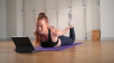 Determined athletic woman exercising, stretching exercises on yoga mat while watching online fitness course video on tablet. Woman practicing yoga online with laptop