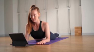 Determined athletic woman exercising, stretching exercises on yoga mat while watching online fitness course video on tablet. Woman practicing yoga online with laptop