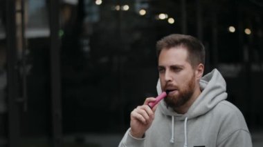 Close-up of a bearded attractive man smoking an electronic cigarette on the street in t evening