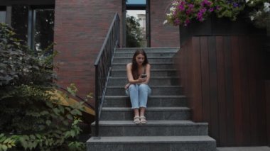 Pretty positive young woman sit on steps outdoors, scroll social media