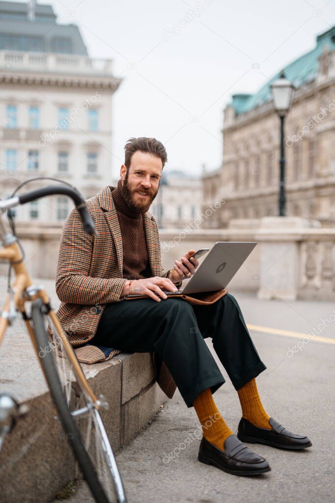 Stylish man sitting outside with laptop and mobile. Online call. Working outside