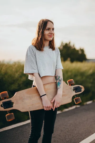 Hauska nuori tyttö farkut ja top seisoo iso longboard. Vihreä luonto — kuvapankkivalokuva