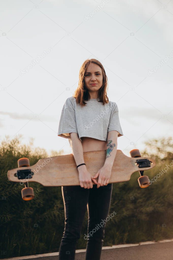 Front view , low angle .Funny young girl in jeans and top stands with big longboard. Green nature background