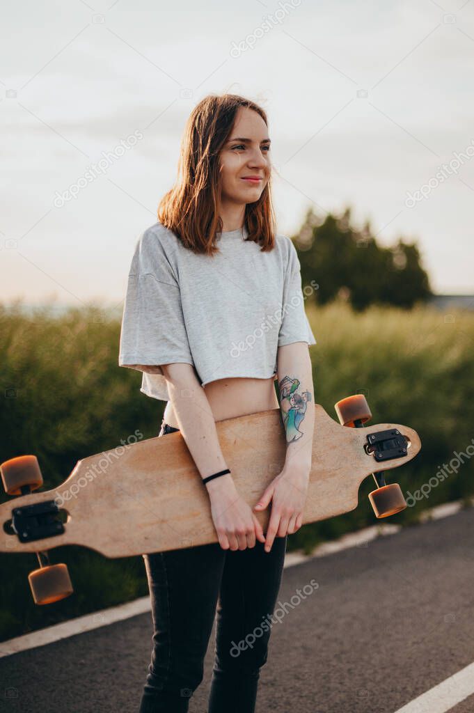 Funny young girl in jeans and top stands with big longboard. Green nature background