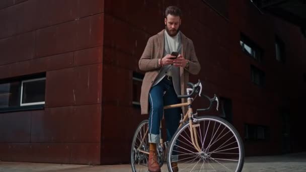 Jeune homme avec vélo tenant un smartphone devant le bâtiment. Homme d'affaires créatif dans un secteur d'activité moderne. — Video