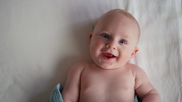 Pequeno menino causiano bonito com olhos azuis sorrindo e deitado na cama no quarto. Com espaço de cópia. Olhando para a câmera. — Vídeo de Stock