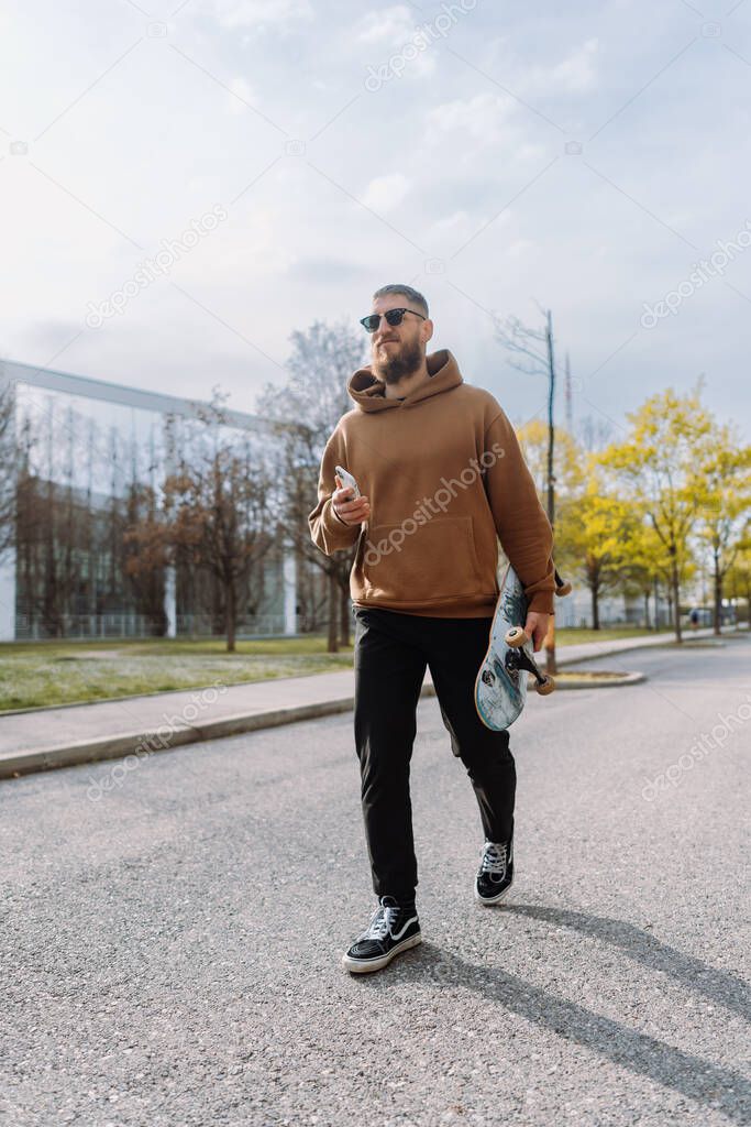 Young stylish man with beard holds a skateboard and smartphone. Vertical