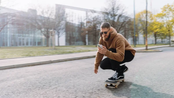 Beau homme monte sur un skateboard et regarde dans son smartphone. espace de copie. — Photo