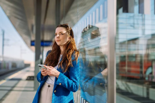Ung kvinne venter på togstasjonen. Holder smarttelefonen og venter på toget. Offentlig transport. – stockfoto