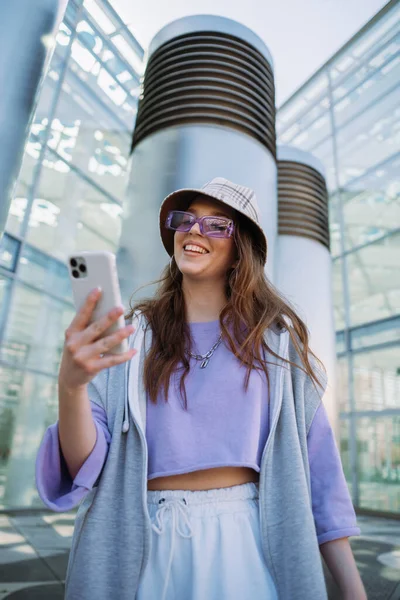 Beautiful young woman in sunglasses and oversize clothes holding modern smartphone. She looking away and smiling — kuvapankkivalokuva