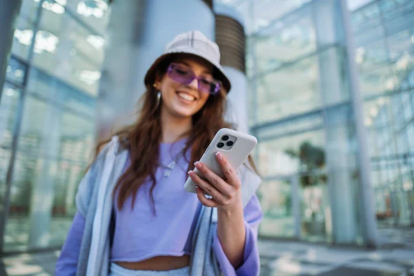 Ung, hvit jente smiler glad med smarttelefonen i byen. – stockfoto