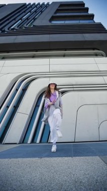 Hip hop kıyafetli neşeli beyaz kız sokak altında dans ederken müzik dinliyor. Dikey video