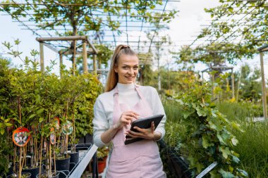 Elinde tablet tutan kadın çiçekçi. Bahçe merkezi sahibi kameraya gülümsüyor..