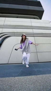 Kulaklıklı mutlu genç bir kadın şehir sokaklarında dans ediyor ve yalnız eğleniyor. Neşeli, çekici, kaygısız kadın akıllı telefonuyla müzik dinliyor. Dikey görünüm