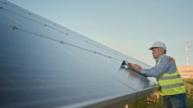 Mature industrial worker using tablet while monitoring rows of photovoltaic solar panels at sunset. Solar park and alternative energy concept