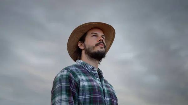 Bearded man wearing plaid shirt holding spikelet, reed, dry grass his mouth and looking at the sky while waiting for the storm at wheat field. Farmer or ranch owner concept