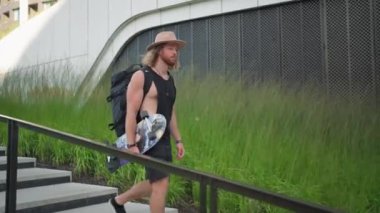 Positive young man walking outdoors by street with skateboard and preparing for the training. Caucasian male person carrying his board down the street