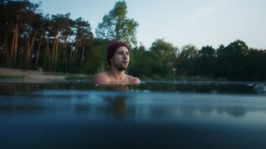 Serious man chills and shakes while standing inside cold winter lake at morning. Forest at the background. Winter swim and cold treatment concept