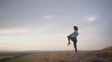 Caucasian female runner standing and warming up in preparation for a jogging run. She is located on a high cliff with beautiful mountains in the background