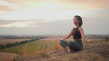 Young beautiful woman doing sport fitness yoga position on a grassy hill during the evening. Girl sitting with closed eyes and keeping prayer gesture