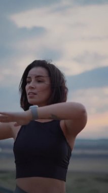 Vertical view of the young sporty woman in sportswear stretching on hill top, warming up and doing twists on cloudy sky background. Fitness, healthy way of life, wellbeing, freedom, loosing weight
