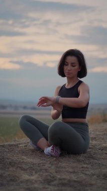 Vertical view of the woman meditating sitting in lotus on the natural background. Girl looking at smartwatch while making breathing exercise. Yoga, meditation, relax and healthy lifestyle concept