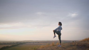 Caucasian female runner standing and warming up in preparation for a jogging run. She is located on a high cliff with beautiful mountains in the background