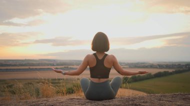 Athletic lady is doing fitness among green wild nature. Woman sitting in the lotus pose and using yoga practices. Relaxing outside concept