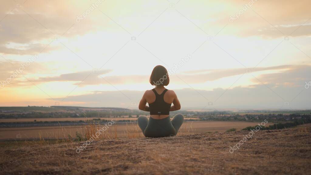 Young beautiful woman doing sport fitness yoga position on a grassy hill during the evening. Girl sitting with closed eyes and keeping prayer gesture