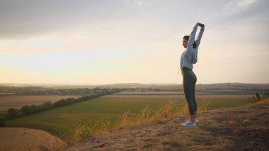 Caucasian female runner standing and warming up in preparation for a jogging run. She is located on a high cliff with beautiful mountains in the background