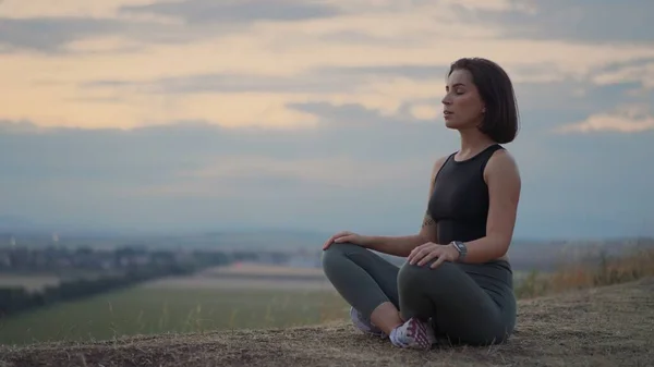 Full length view of the mindful young woman making mudra gesture, sitting in lotus position at the grassy hill. Peaceful millennial girl deeply meditating, doing breathing yoga exercises alone