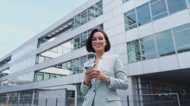 Low angle view of the confident businesswoman using smartphone while going through the street. Happy lady making work call in the urban space